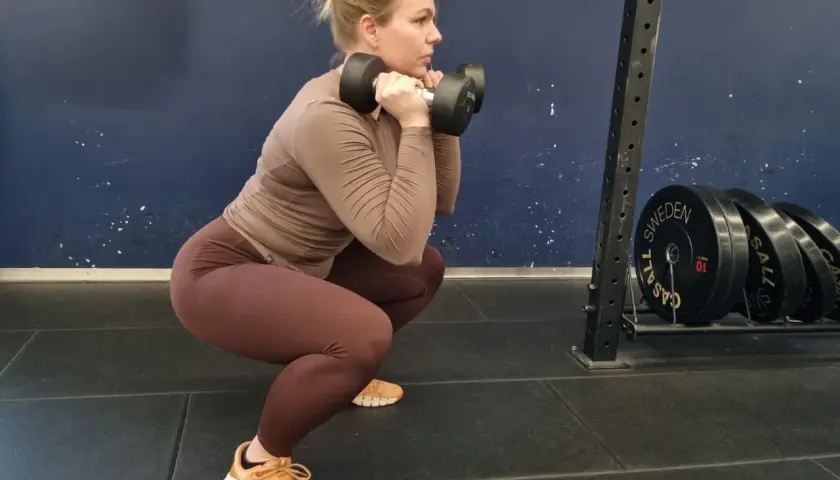 Kvinna gör squats med hantlar framför en mörkblå vägg. Hon har bruna träningskläder och ljusblont hår uppsatt i en knut.