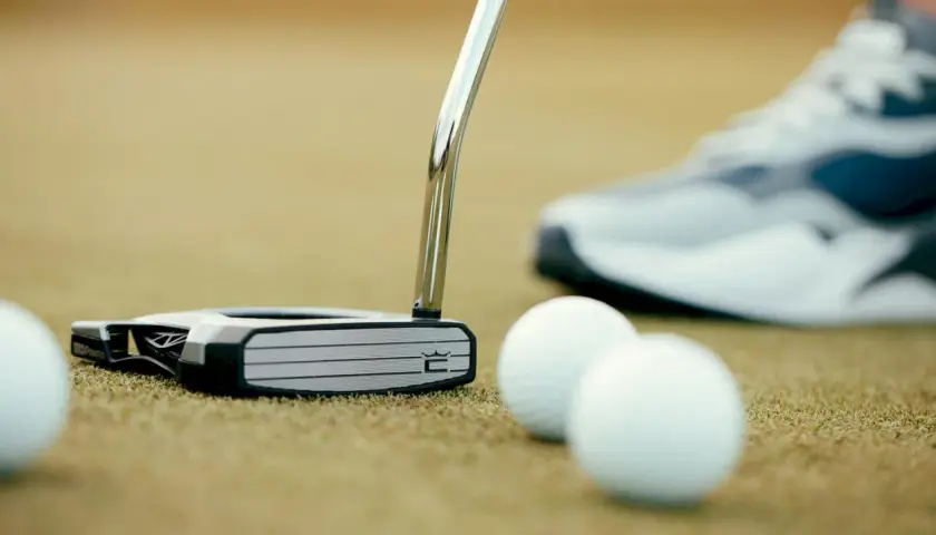 En golfare funderar vilken putter ska man ha. En modern putter ligger på greenen bredvid golfbollar, med en sko i bakgrunden.