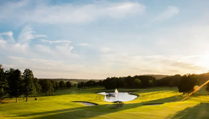 Solnedgång över en golfbana med en damm och fontän. En spelare är på väg att slå ett slag, kanske för en albatross.
