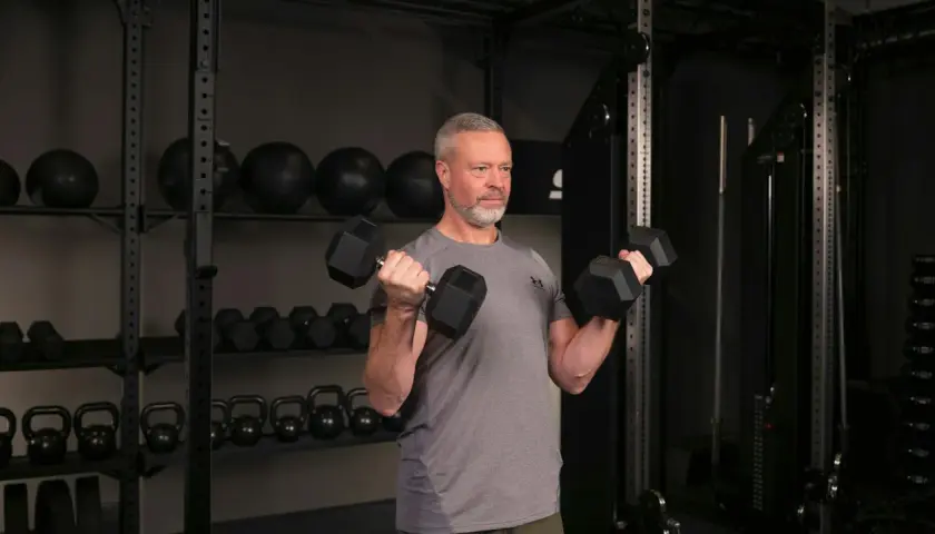 Man med grått hår och skägg utför sittande bicepscurl med hantlar i ett gym.