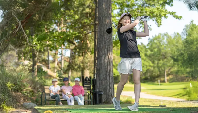 En flicka svingar sin golfklubba på ett soligt golfhål. Tre barn sitter på en bänk i bakgrunden och tittar på.