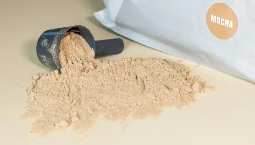 Mörkblå skopa med ljusbrunt pulver, en del av det har spillts ut. Påsen i bakgrunden har en brun etikett med texten "MOCHA". Detta visar vad som kan finnas i proteinpulver.