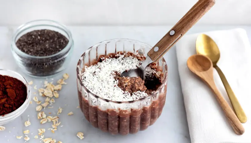 En skål med overnight oats chokladboll, toppad med kokosflingor. Bredvid finns ingredienser som havregryn, chiafrön och kakaopulver.