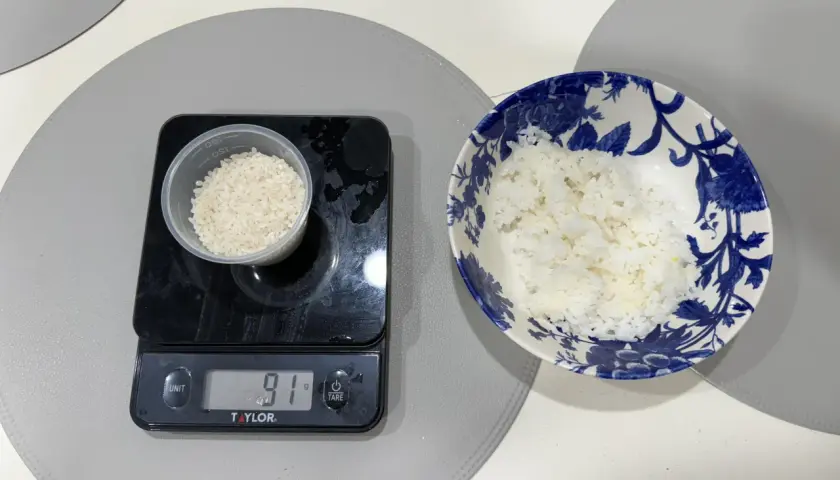 En digitalvåg visar 91 gram ris i en liten behållare. Bredvid står en skål med mer ris, redo att ätas. Perfekt för att hålla koll på råris kalorier.