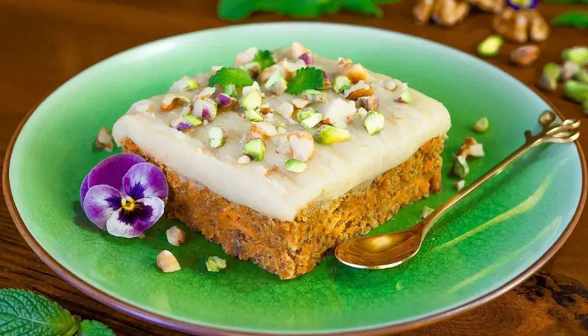 En bit sockerfri morotskaka med krämig frosting, pistagenötter och mynta på en grön tallrik. En viol och en guldig sked fulländar presentationen.