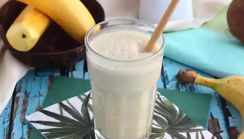 En krämig banan milkshake med en bambustrå i ett glas, omgiven av bananer och en kokosnöt.