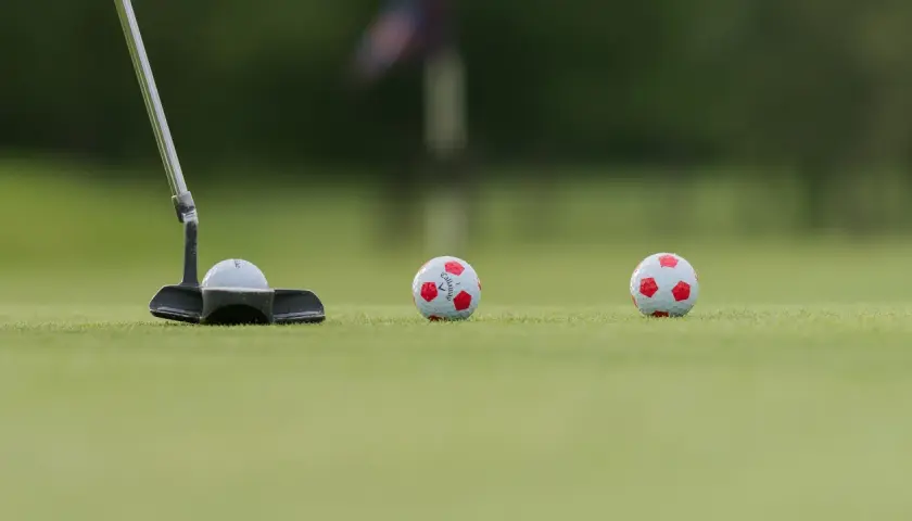 En golfputter är redo att slå en boll med fotbollsmönster. Två likadana bollar ligger längre bort på greenen.