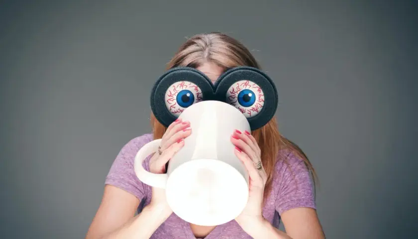 Kvinna med stora, uppspärrade ögon och en mugg framför ansiktet, en bild av koffeinberoende.
