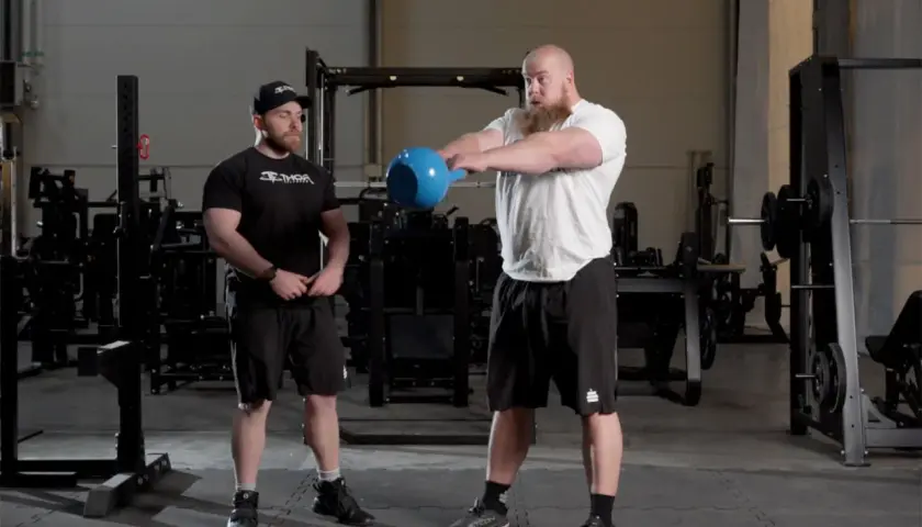 Man med skägg utför kettlebell swing varje dag i gymmet. Bredvid står en tränare.