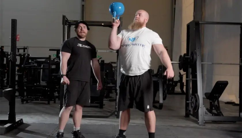 Man med skägg utför en kettlebell press med en blå kettlebell. En annan man står bredvid och tittar.