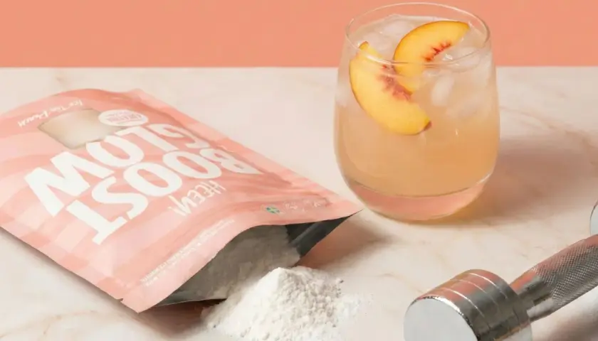 En påse "Glow Boost" med persikosmak, en drink med persika, och en hantel. Frågan "hur mycket kreatin per dag" kan besvaras med denna produkt.