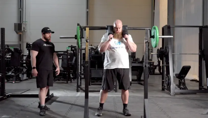 Man med skägg förbereder sig för knäböj med en safety squat bar. En tränare står bredvid.