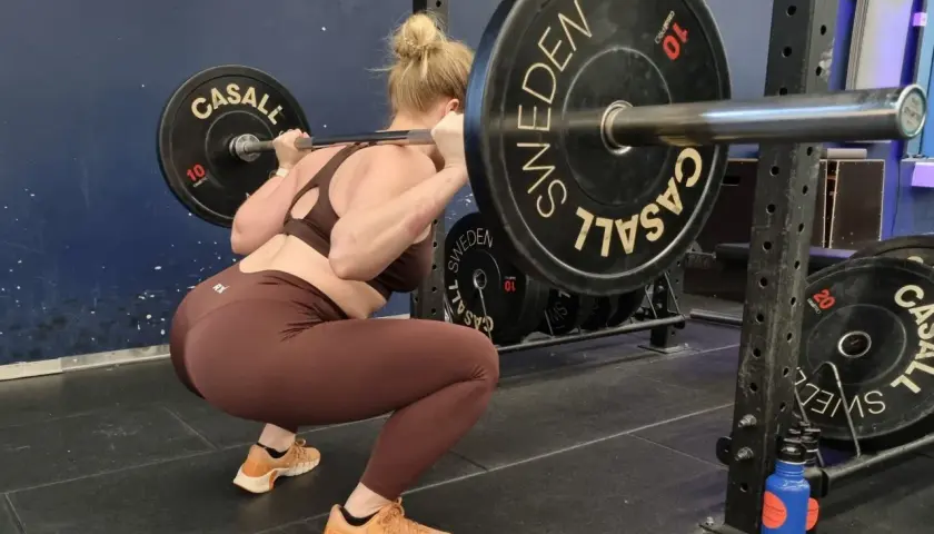 Kvinna utför squats med vikt. Stången vilar på axlarna, med tunga viktskivor märkta "SWEDEN" och "CASALL".