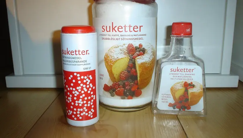 Tre förpackningar Suketter, ett sockerersättningsmedel, står på ett träbord. En burk, en tub och en flaska. Suketter är utmärkt till bakning och matlagning, helt utan kalorier.