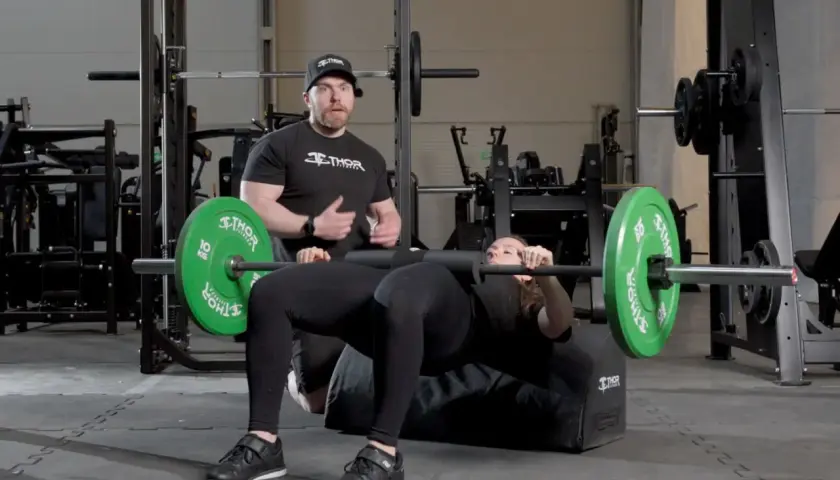 Man instruerar kvinna som utför hip thrust med skivstång på en bänk i gym.