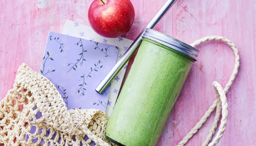 En grön, proteinrik smoothie i glasburk med metallrör, bredvid ett rött äpple och en vävd kasse på en rosa bakgrund.