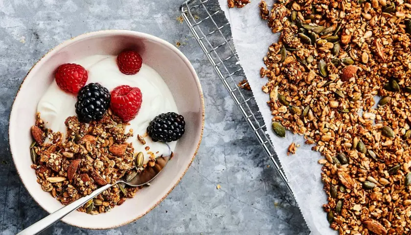 En skål med yoghurt, bär och hemgjord granola – en perfekt frukost utan kolhydrater. Granola med nötter och frön ligger bredvid på ett bakplåtspapper.