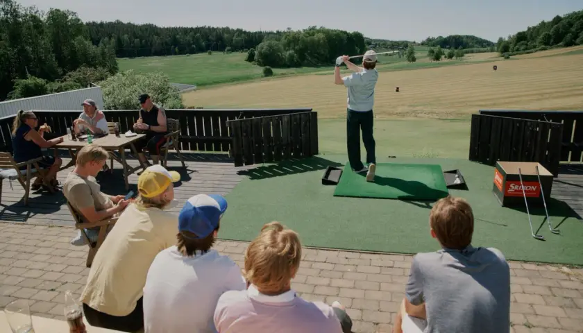 En solig dag på golfbanan. En person slår ett slag medan andra sitter och tittar på, kanske diskuterar annorlunda spelformer golf.