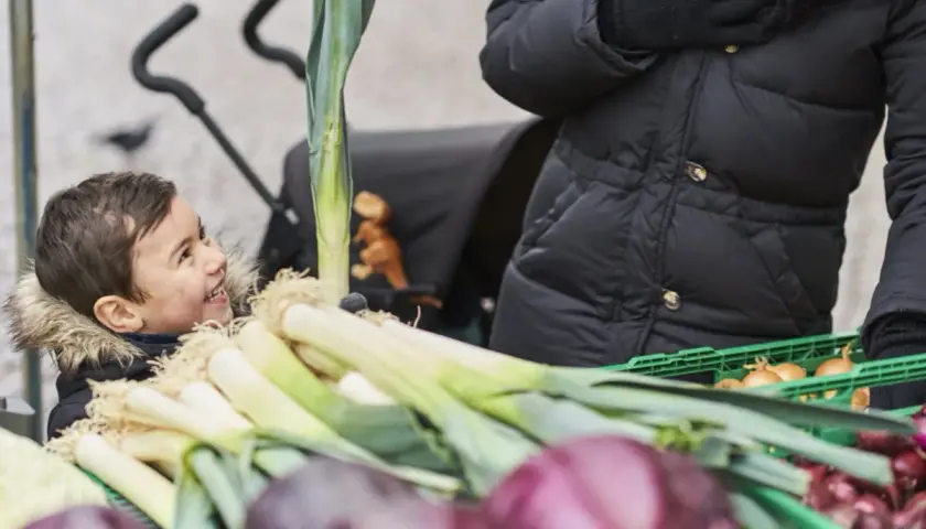 Glad pojke tittar på purjolök och lök på en marknad. En person i svart jacka håller i en grön låda med billiga råvaror.