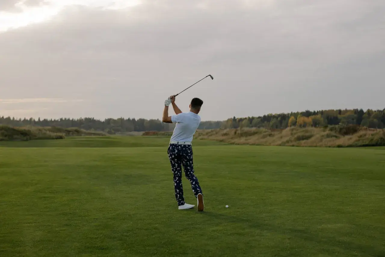 En man svingar sin golfklubba på en grön golfbana. Frågan "hur lång tid tar en golfrunda" kan besvaras med att det beror på många faktorer.