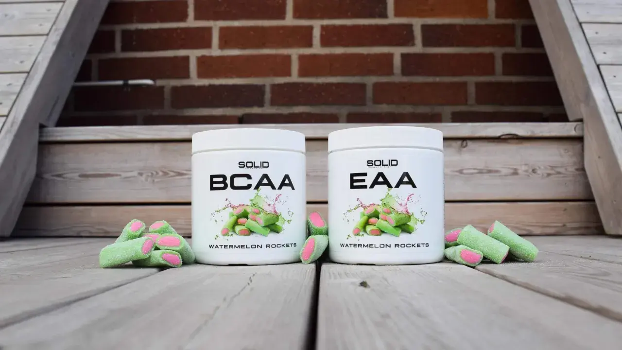 Två burkar, en med BCAA och en med EAA, står framför en tegelvägg. Gröna godisar med rosa inre ligger utspridda.