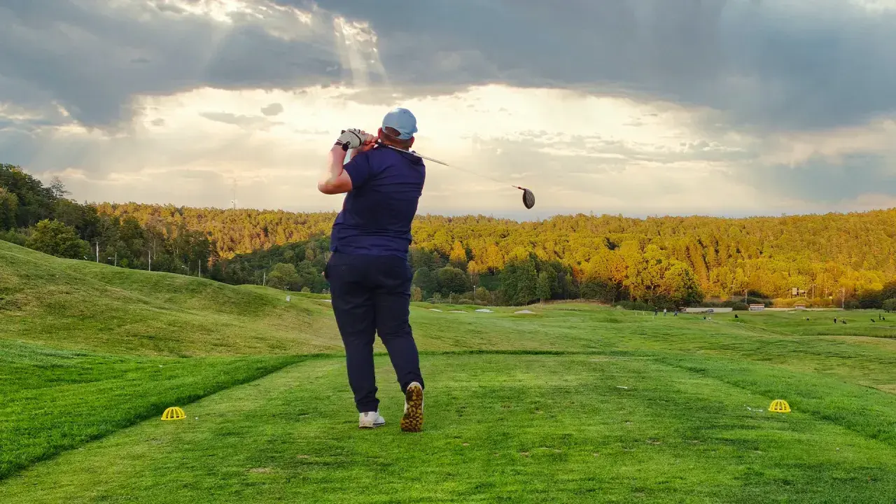 En golfare svingar klubban på en utslagsplats i golf. Solstrålar bryter igenom molnen över en grönskande bana.
