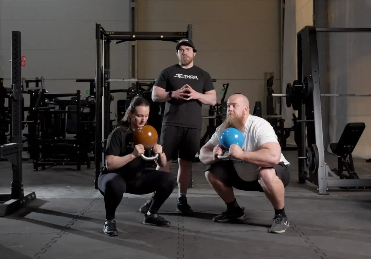Två personer utför goblet squats med kettlebells, en instruktör står bakom.