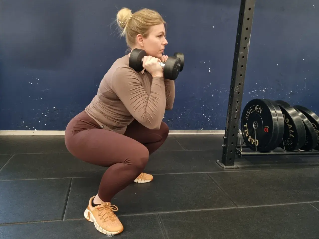 Kvinna gör squats med hantlar framför en mörkblå vägg. Hon har bruna träningskläder och ljusblont hår uppsatt i en knut.