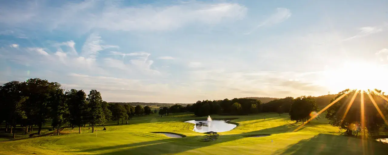 Solnedgång över en golfbana med en damm och fontän. En spelare är på väg att slå ett slag, kanske för en albatross.