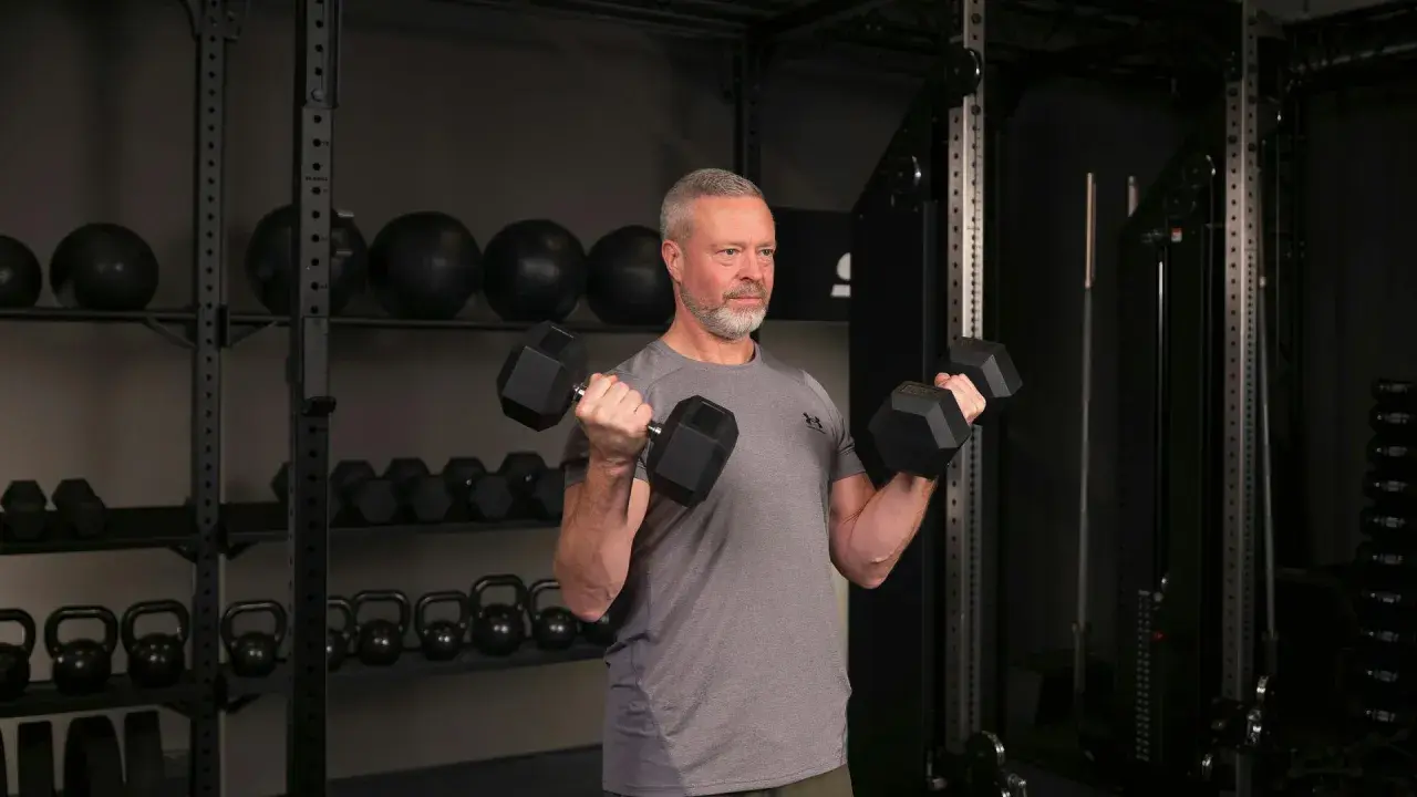 Man med grått hår och skägg utför sittande bicepscurl med hantlar i ett gym.