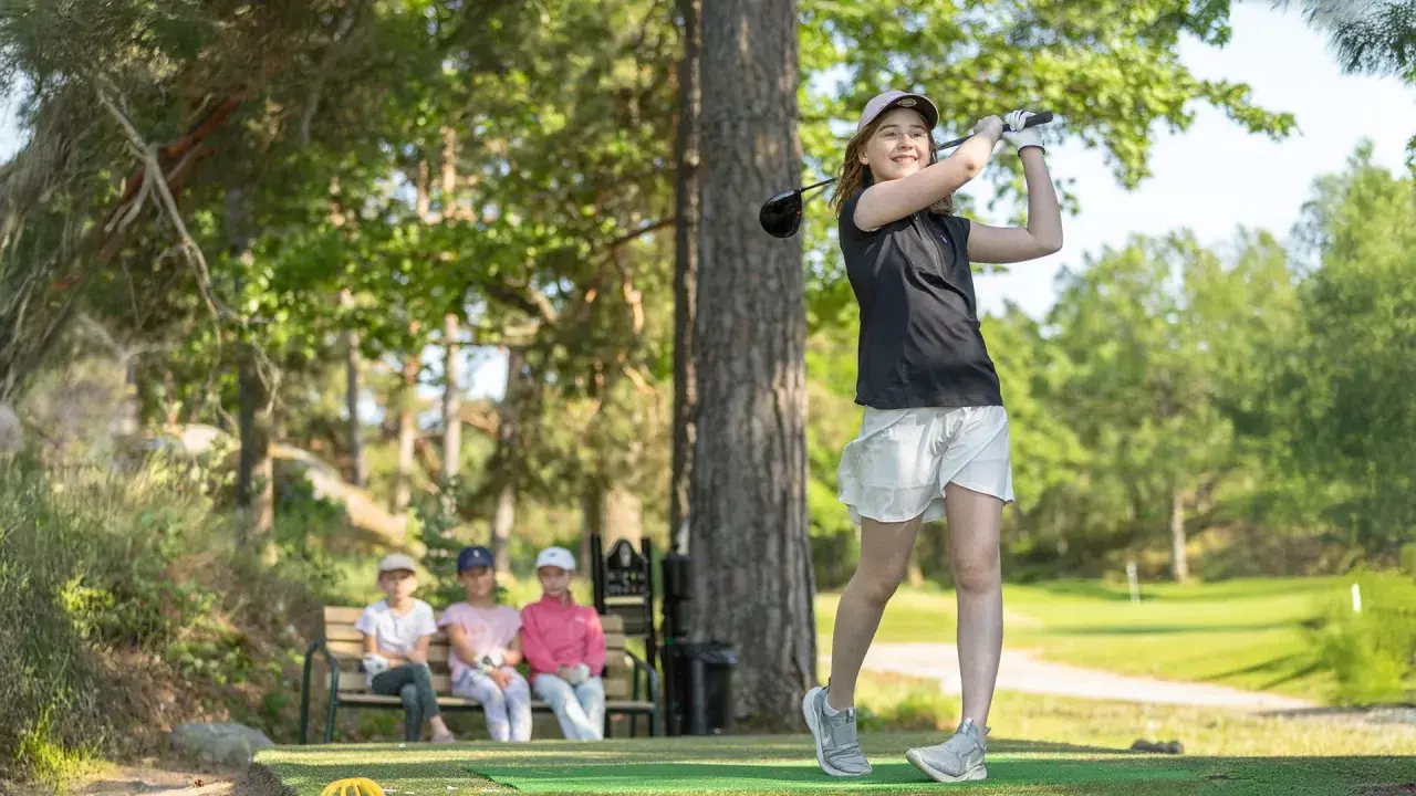 En flicka svingar sin golfklubba på ett soligt golfhål. Tre barn sitter på en bänk i bakgrunden och tittar på.