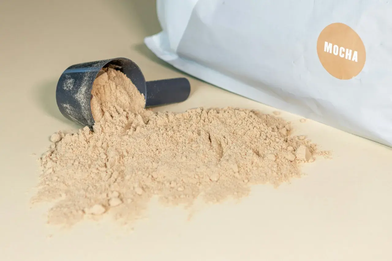 Mörkblå skopa med ljusbrunt pulver, en del av det har spillts ut. Påsen i bakgrunden har en brun etikett med texten "MOCHA". Detta visar vad som kan finnas i proteinpulver.
