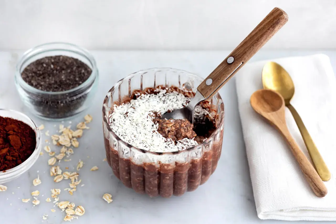En skål med overnight oats chokladboll, toppad med kokosflingor. Bredvid finns ingredienser som havregryn, chiafrön och kakaopulver.