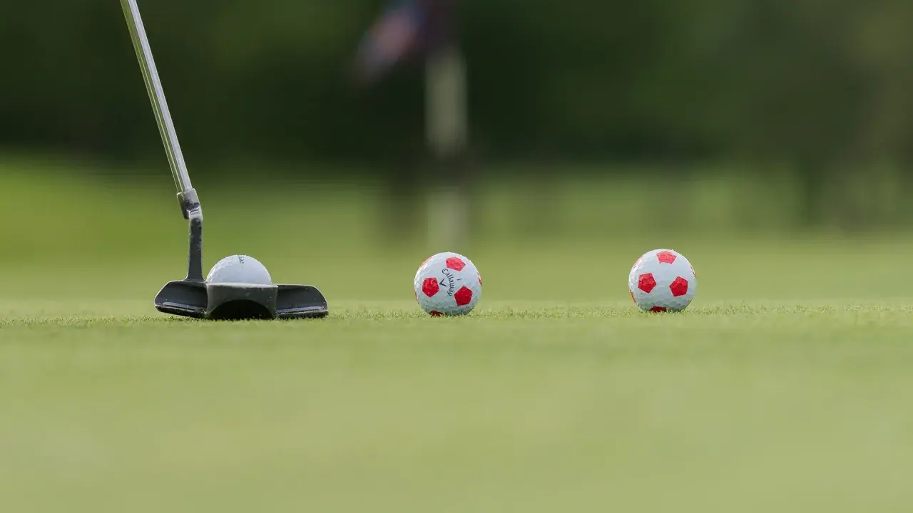 En golfputter är redo att slå en boll med fotbollsmönster. Två likadana bollar ligger längre bort på greenen.