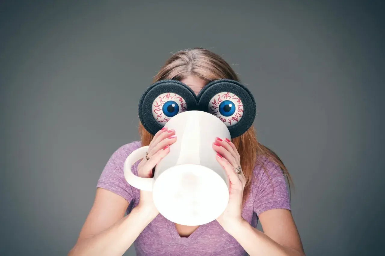 Kvinna med stora, uppspärrade ögon och en mugg framför ansiktet, en bild av koffeinberoende.