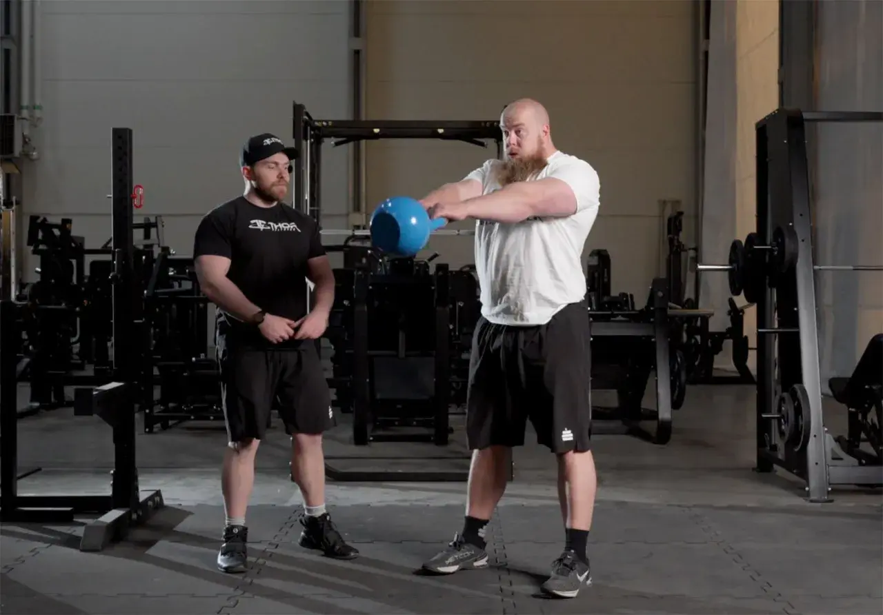 Man med skägg utför kettlebell swing varje dag i gymmet. Bredvid står en tränare.