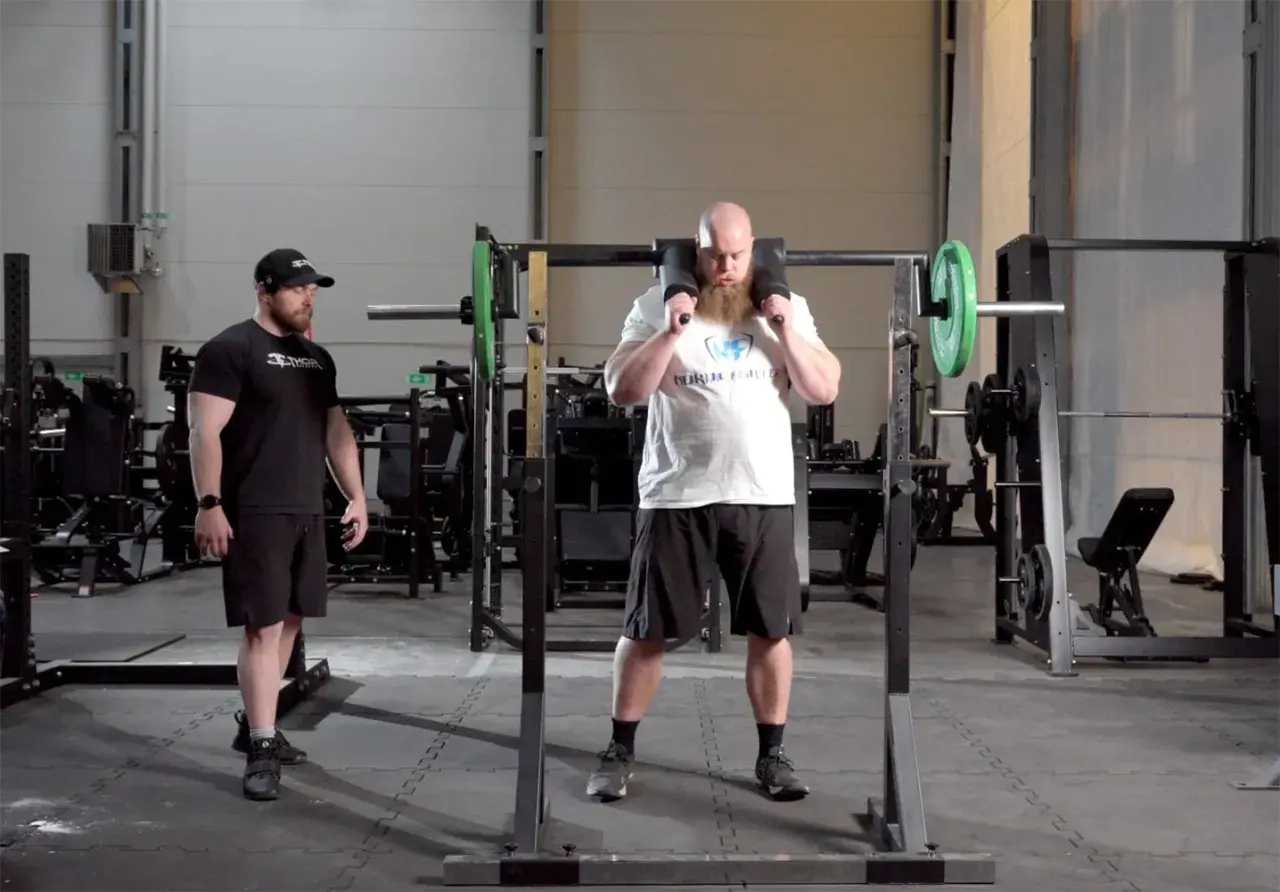 Man med skägg förbereder sig för knäböj med en safety squat bar. En tränare står bredvid.