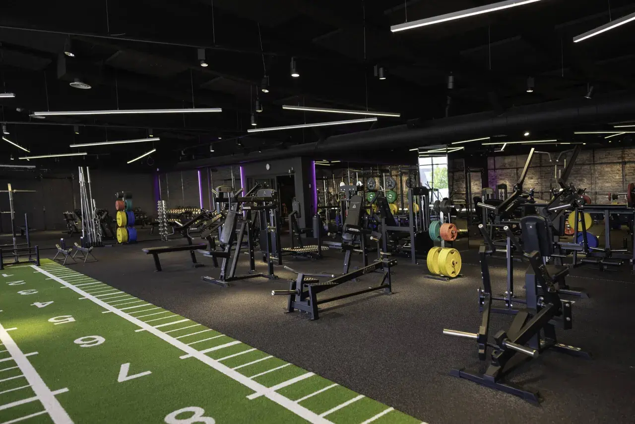 Gym med moderna träningsmaskiner, inklusive en t bar row. Grön konstgräsmatta med markeringar löper längs golvet.