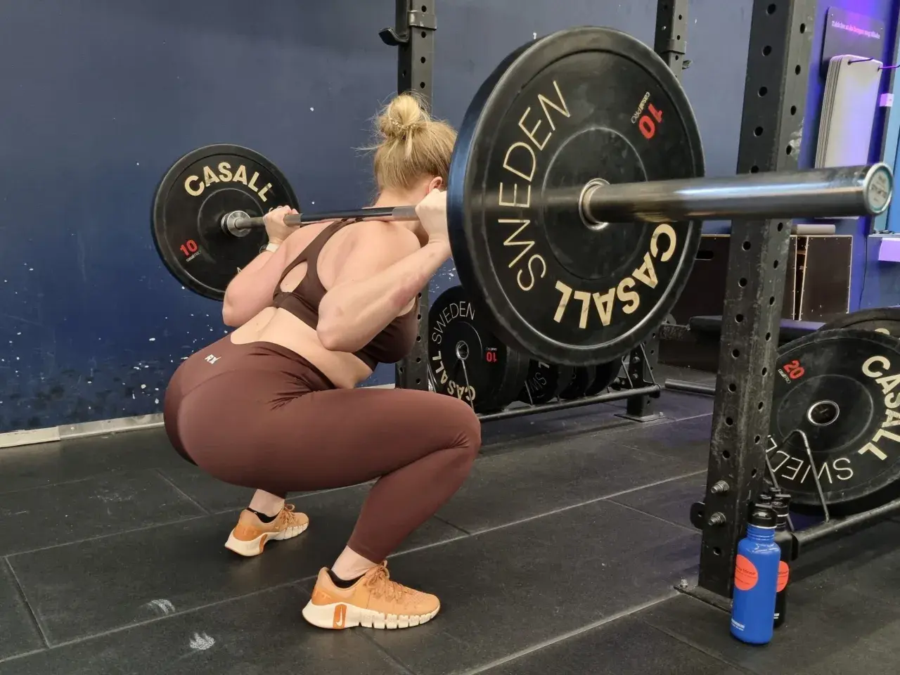 Kvinna utför squats med vikt. Stången vilar på axlarna, med tunga viktskivor märkta "SWEDEN" och "CASALL".