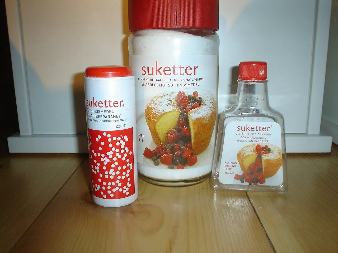 Tre förpackningar Suketter, ett sockerersättningsmedel, står på ett träbord. En burk, en tub och en flaska. Suketter är utmärkt till bakning och matlagning, helt utan kalorier.