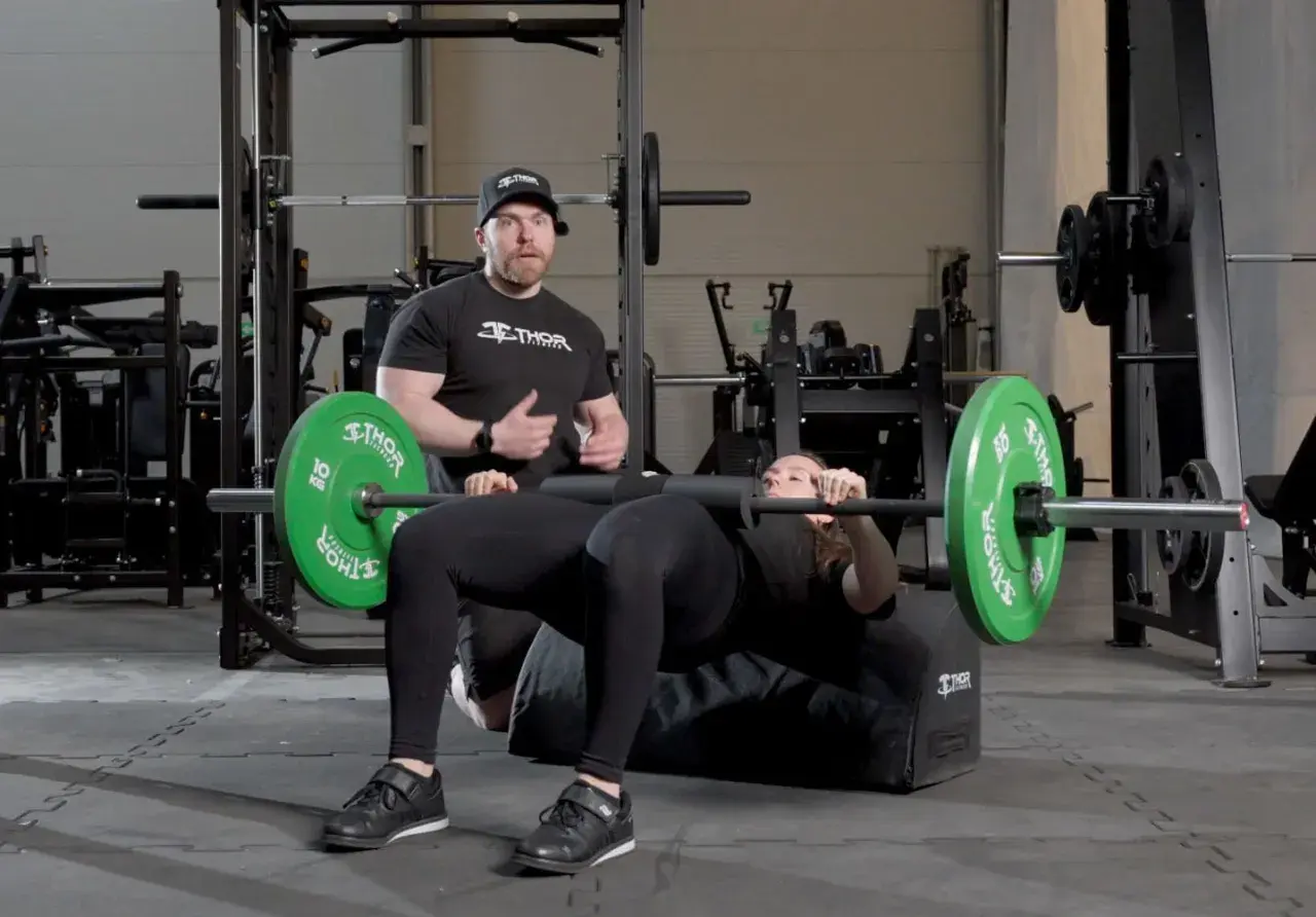 Man instruerar kvinna som utför hip thrust med skivstång på en bänk i gym.