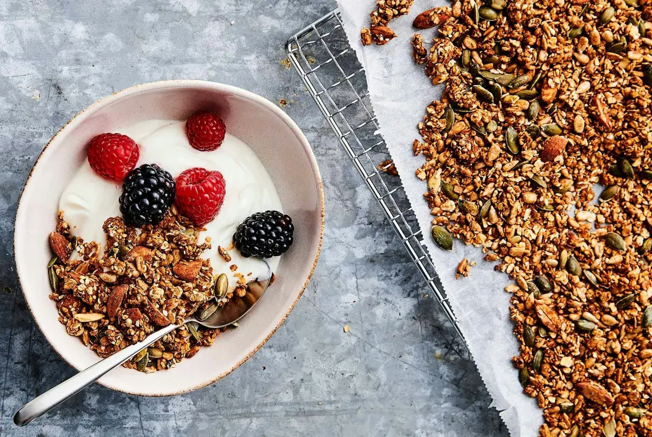 En skål med yoghurt, bär och hemgjord granola – en perfekt frukost utan kolhydrater. Granola med nötter och frön ligger bredvid på ett bakplåtspapper.