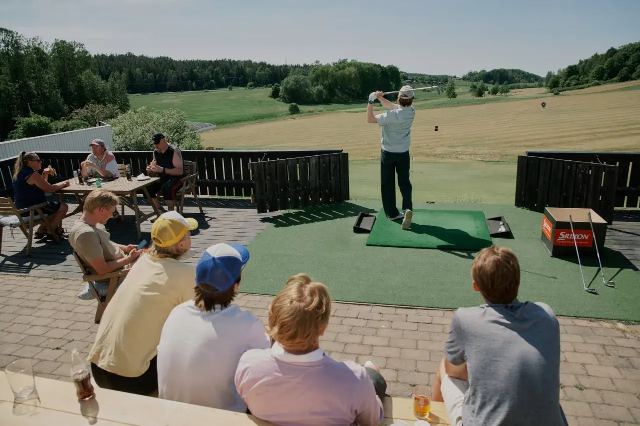 En solig dag på golfbanan. En person slår ett slag medan andra sitter och tittar på, kanske diskuterar annorlunda spelformer golf.