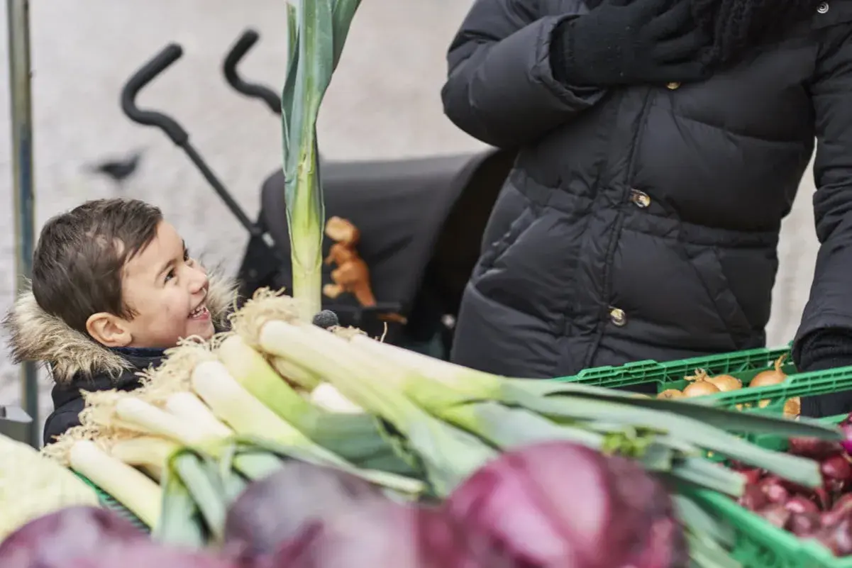 Glad pojke tittar på purjolök och lök på en marknad. En person i svart jacka håller i en grön låda med billiga råvaror.