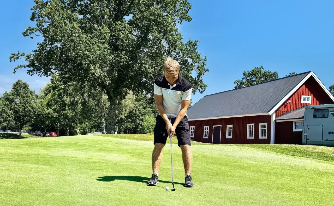 Man puttar på golfbana, perfekt för friskvårdsbidrag golf. Röd byggnad i bakgrunden.