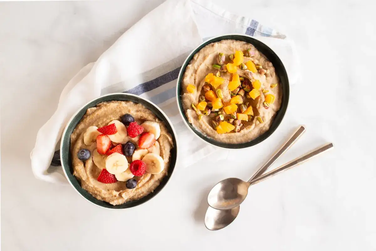 Två skålar med krämigt risgröt, en toppad med bär och banan, den andra med aprikoser och pistagenötter. Ett perfekt recept på cream of rice för en god frukost.
