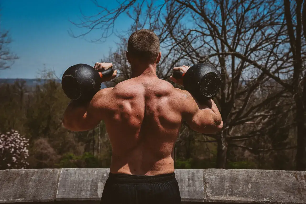 Muskulös man utför kettlebell press utomhus. Tränar axlar och rygg med två tunga vikter.