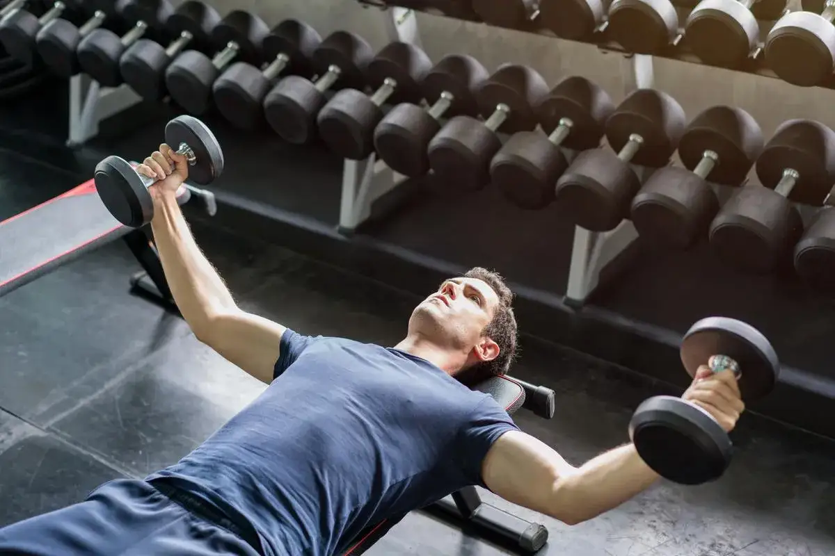 Man tränar bröstmuskler med hantlar på en bänk i ett gym. Hantlar ligger uppradade i bakgrunden.