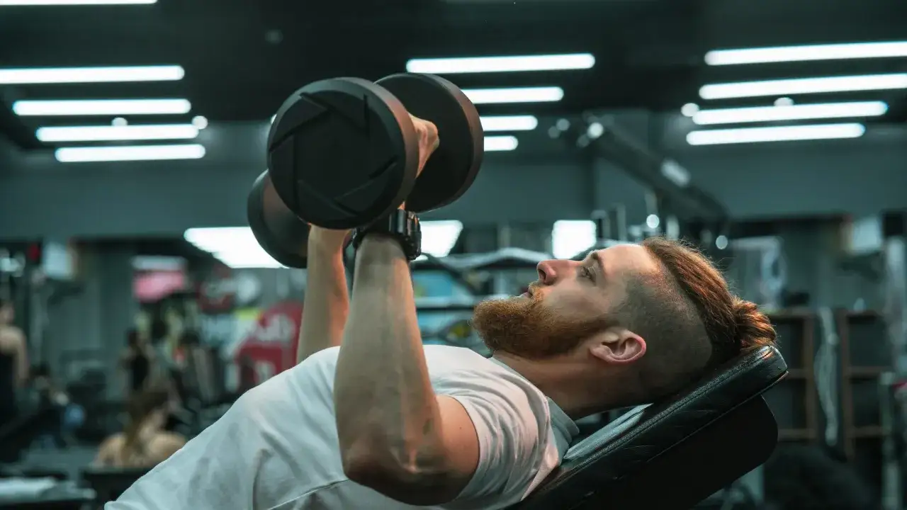 Man med skägg utför lutande hantelpress på gym.