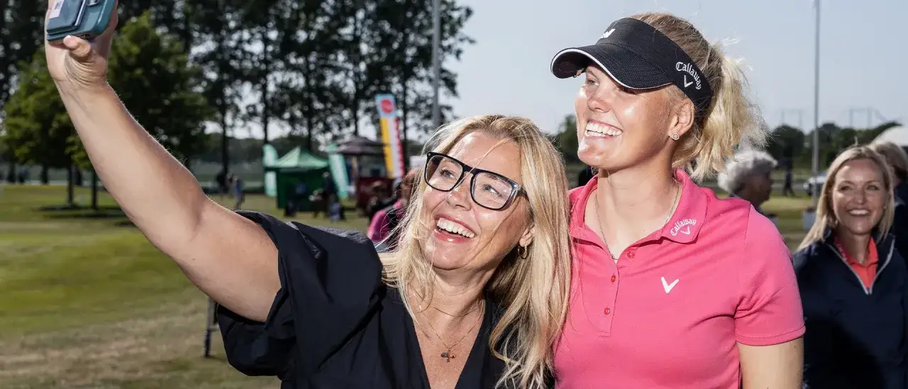 Två glada kvinnor tar en selfie vid Helsingborg Open. En kvinna med glasögon och en golfare i rosa tröja ler mot kameran.
